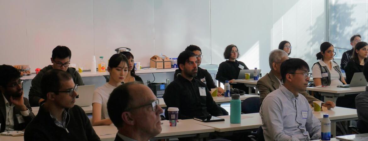 A group of faculty and researchers sit at long white tables in a brightly lit room during the WCGEC Energy Systems Research Workshop. The attendees are focused on a presentation (off-camera), with laptops, notebooks, and coffee cups on the tables in front of them