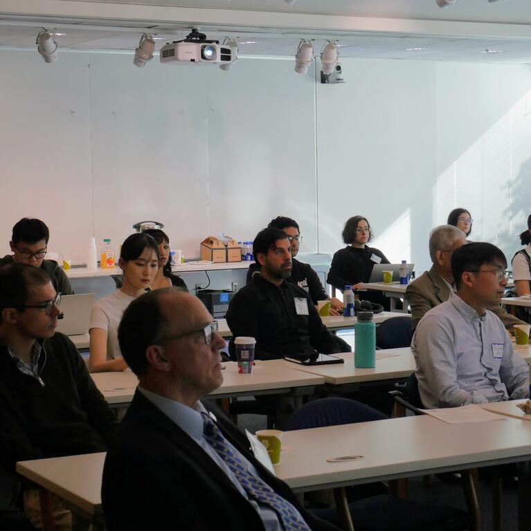 A group of faculty and researchers sit at long white tables in a brightly lit room during the WCGEC Energy Systems Research Workshop. The attendees are focused on a presentation (off-camera), with laptops, notebooks, and coffee cups on the tables in front of them