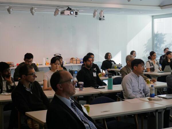 A group of faculty and researchers sit at long white tables in a brightly lit room during the WCGEC Energy Systems Research Workshop. The attendees are focused on a presentation (off-camera), with laptops, notebooks, and coffee cups on the tables in front of them