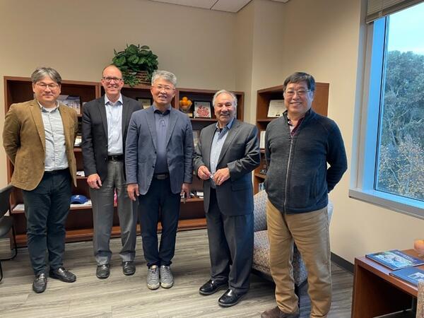 Five men are standing together in an office, smiling at the camera. They are dressed in business casual attire, and behind them is a bookshelf with various items, including plants and books.